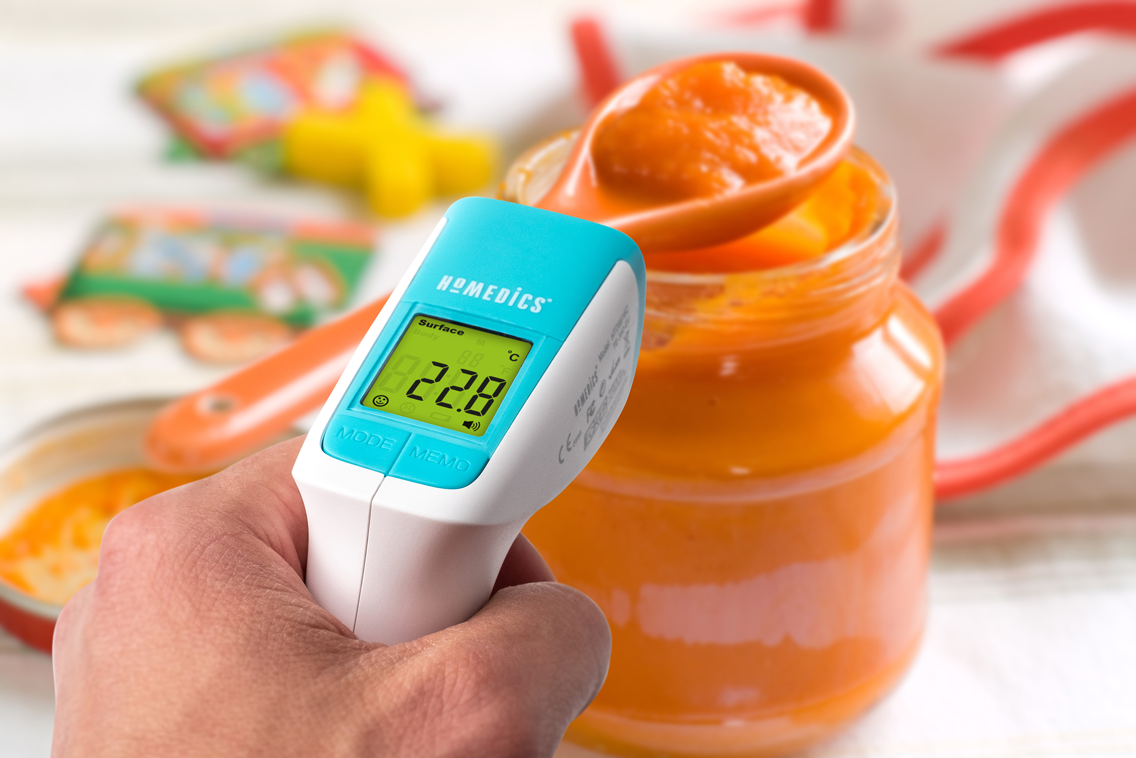 A person holding a thermometer measuring baby food temperature. Jar of baby food in background.