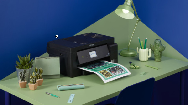 A black Epson printer printing a document on a green desk with a lamp, cactus, and stationary.