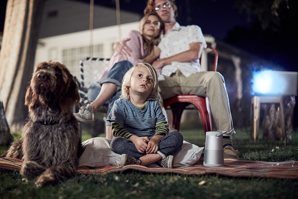 Family watching a movie outdoors. Boy in front, parents on swing, dog on the left, projector on the right.