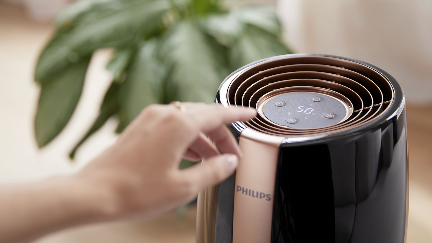 Hand van een persoon raakt een zwarte en bronzen Philips luchtreiniger aan. Een groene plant op de achtergrond.