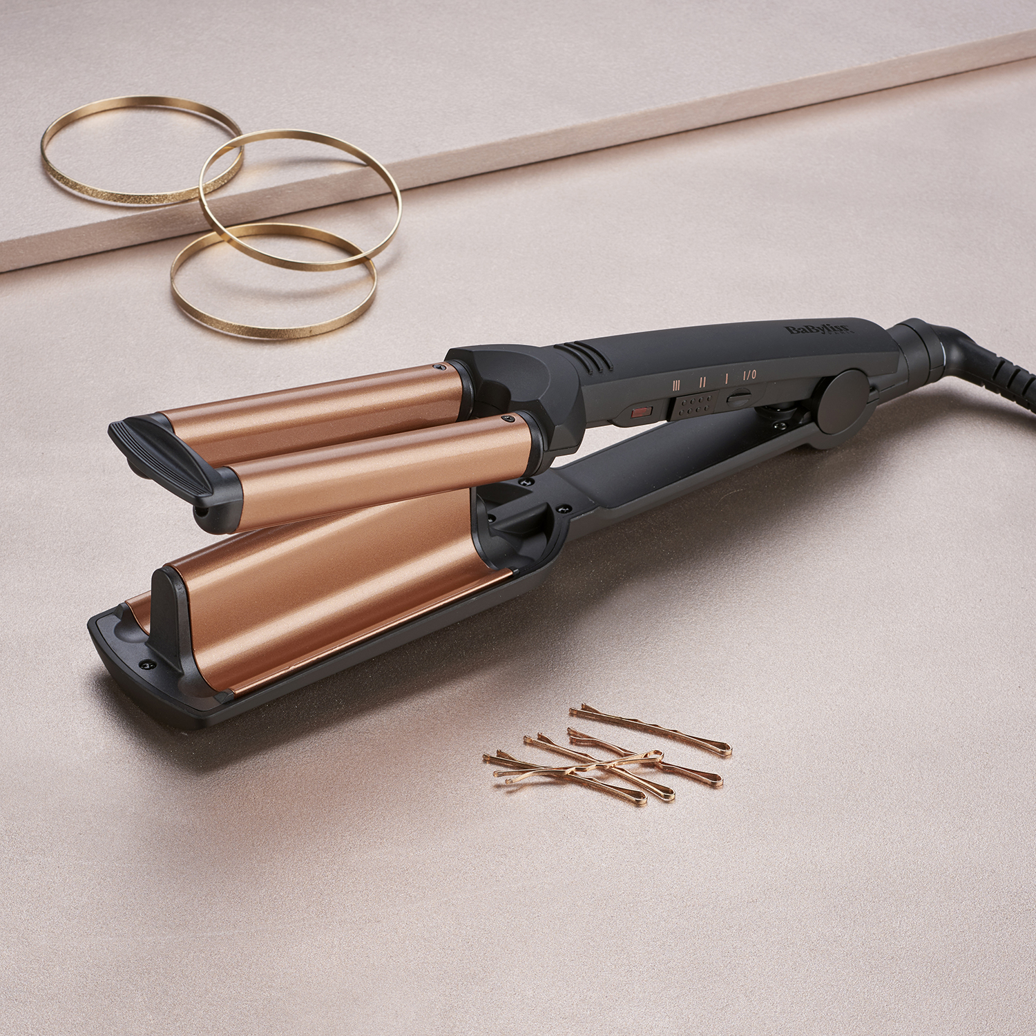 Een Babyliss haarwaver, haarspelden en gouden armbanden op een roze oppervlak.