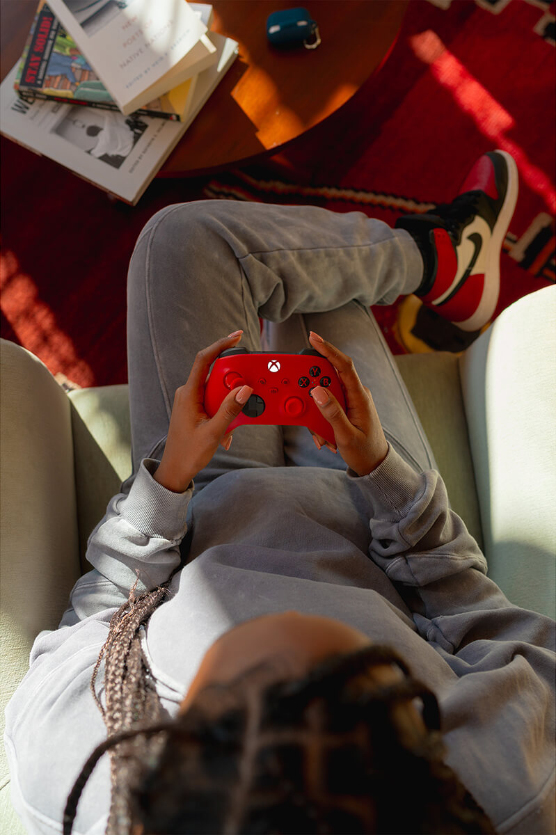 Person lying on a couch, playing a red Xbox controller. The scene is from a high angle.