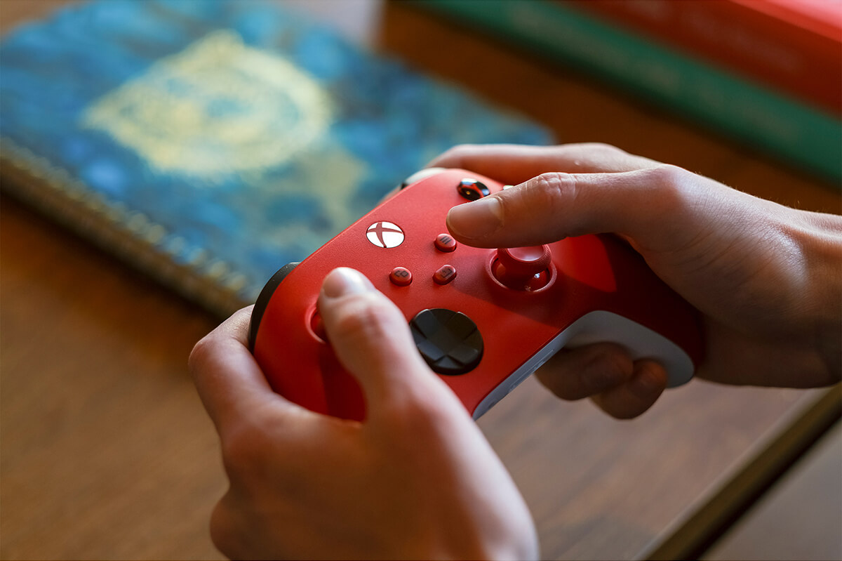 Hands holding a red Xbox controller. Buttons are black and red, with a lit central white X.