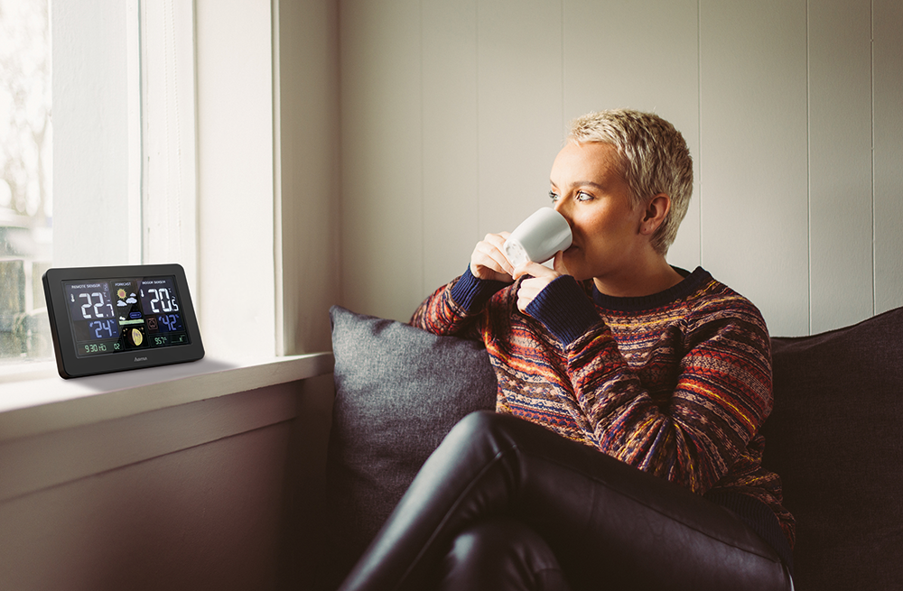 A woman with short hair drinks from a mug while looking at a weather station.