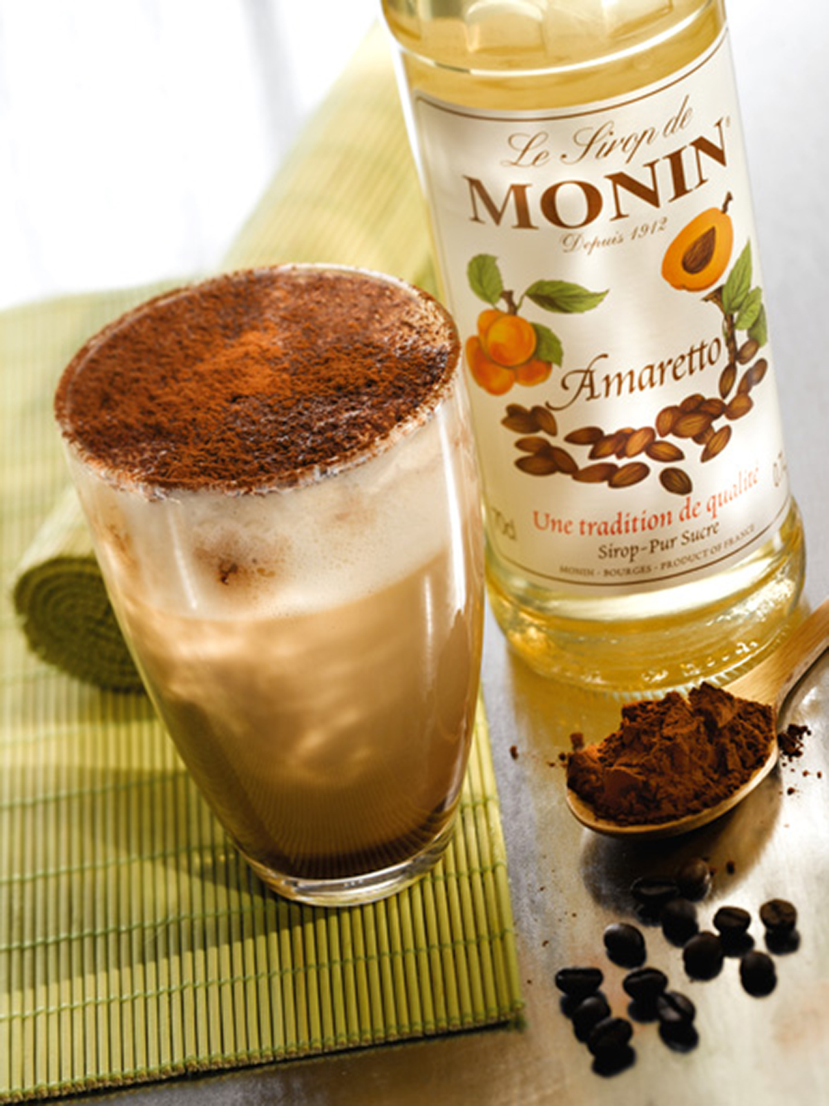 Ein Latte, mit Kakao bestreut, steht neben einer Amaretto-Flasche und Kaffeebohnen.