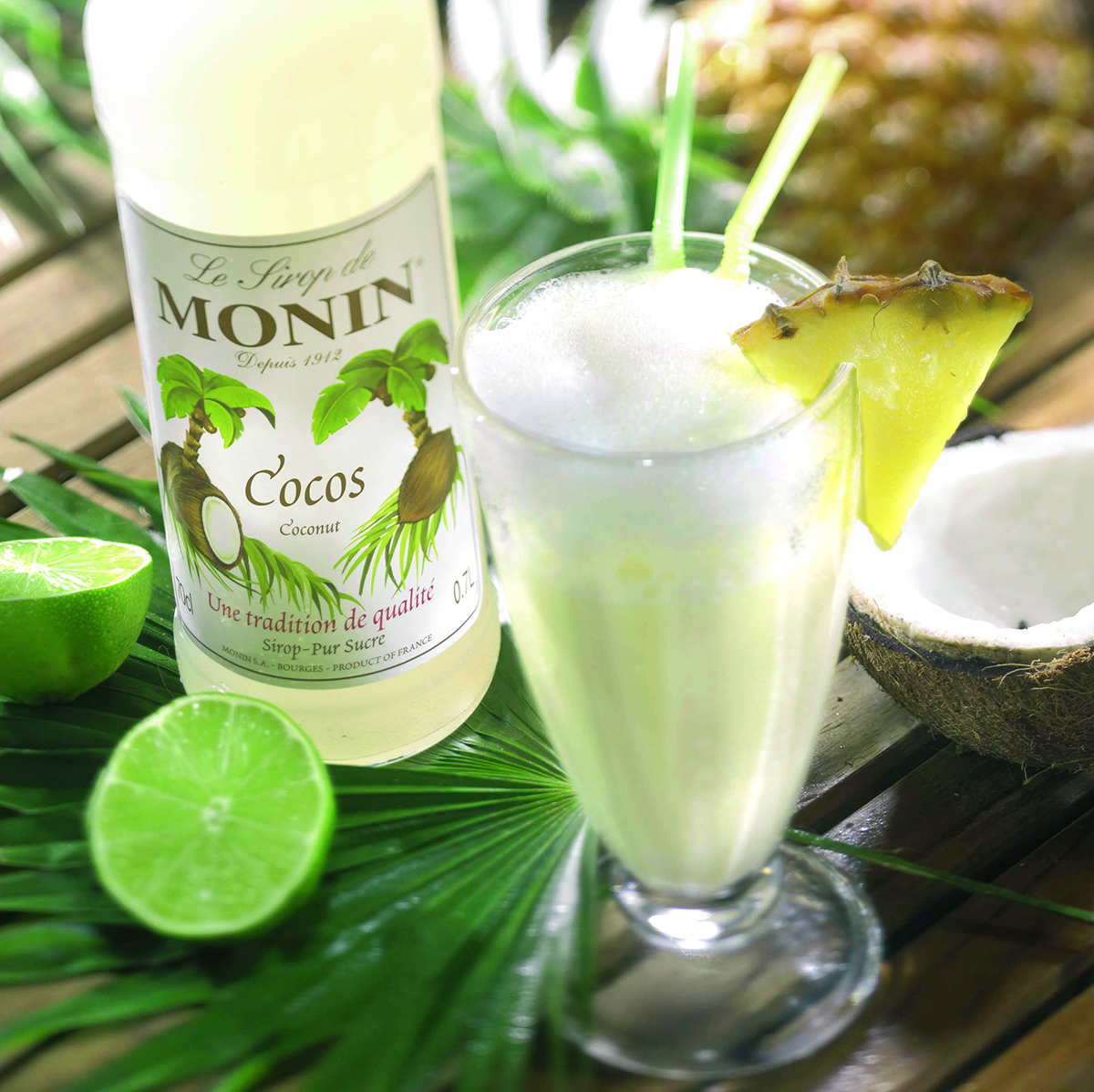 Cocktail mit Ananasscheibe und Limette, neben Kokosnuss. Monin-Sirupflasche.