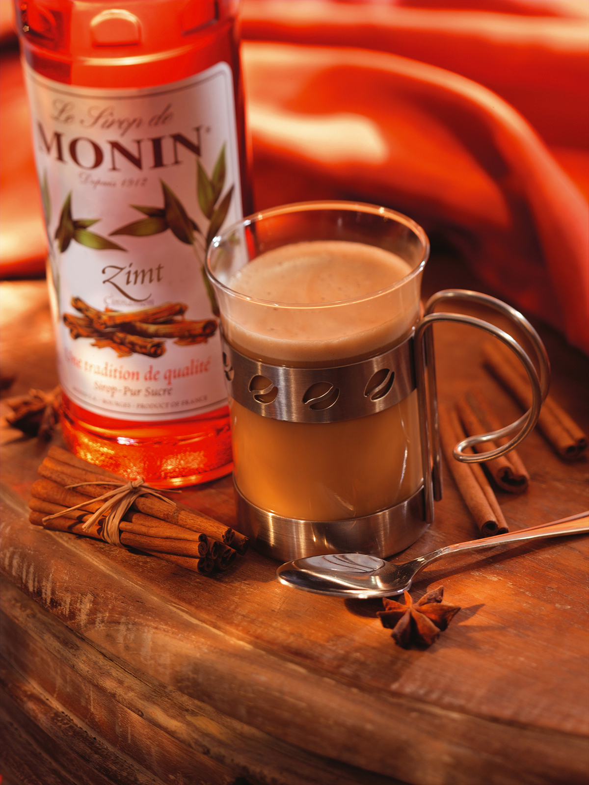Tasse mit Getränk, Monin-Sirupflasche, Zimtstangen und Sternanis auf Holzoberfläche.