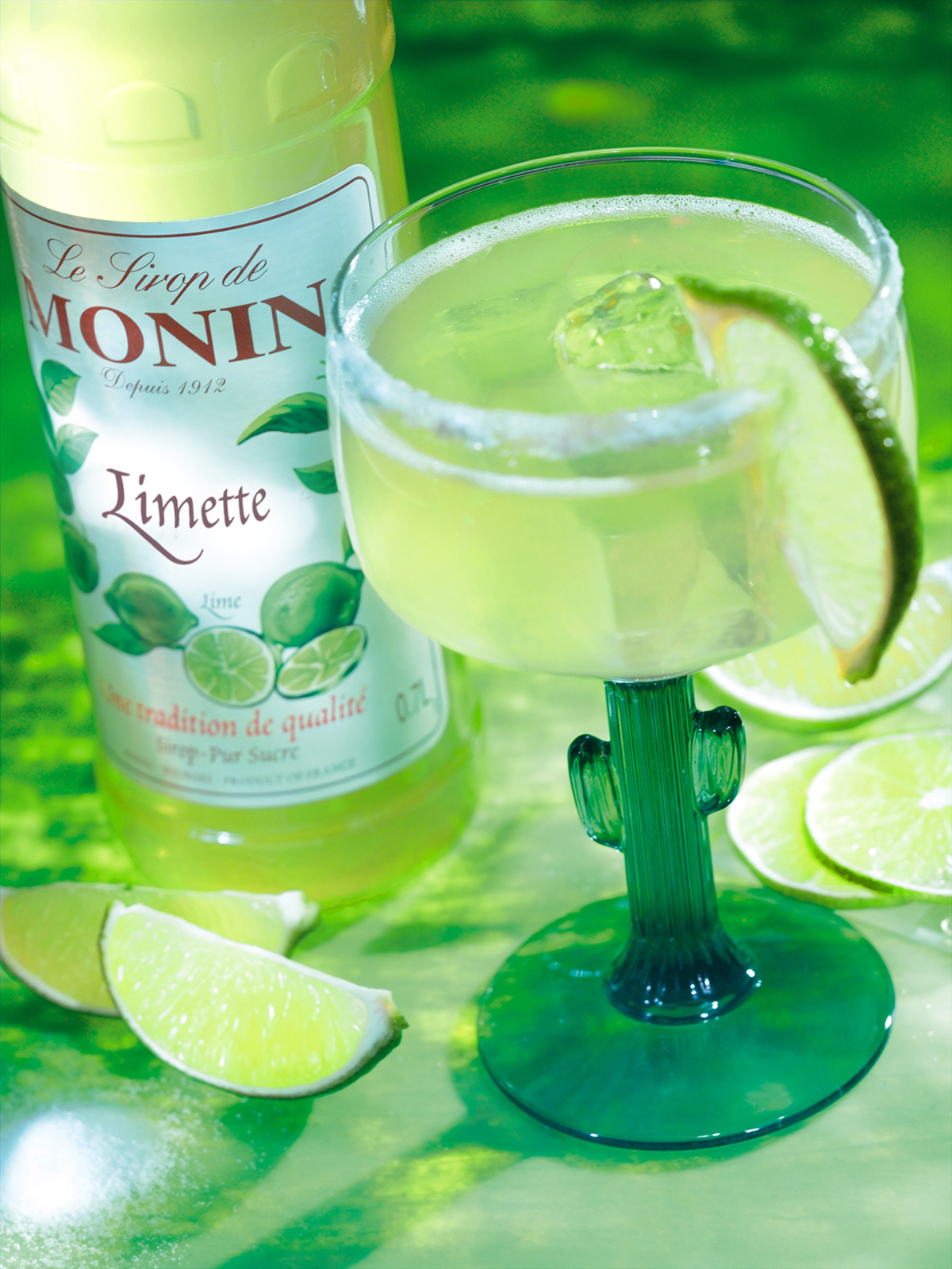Ein Limetten-Margarita mit Monin-Sirup und Limettenscheiben auf grüner Oberfläche, mit einem Kaktusglas.