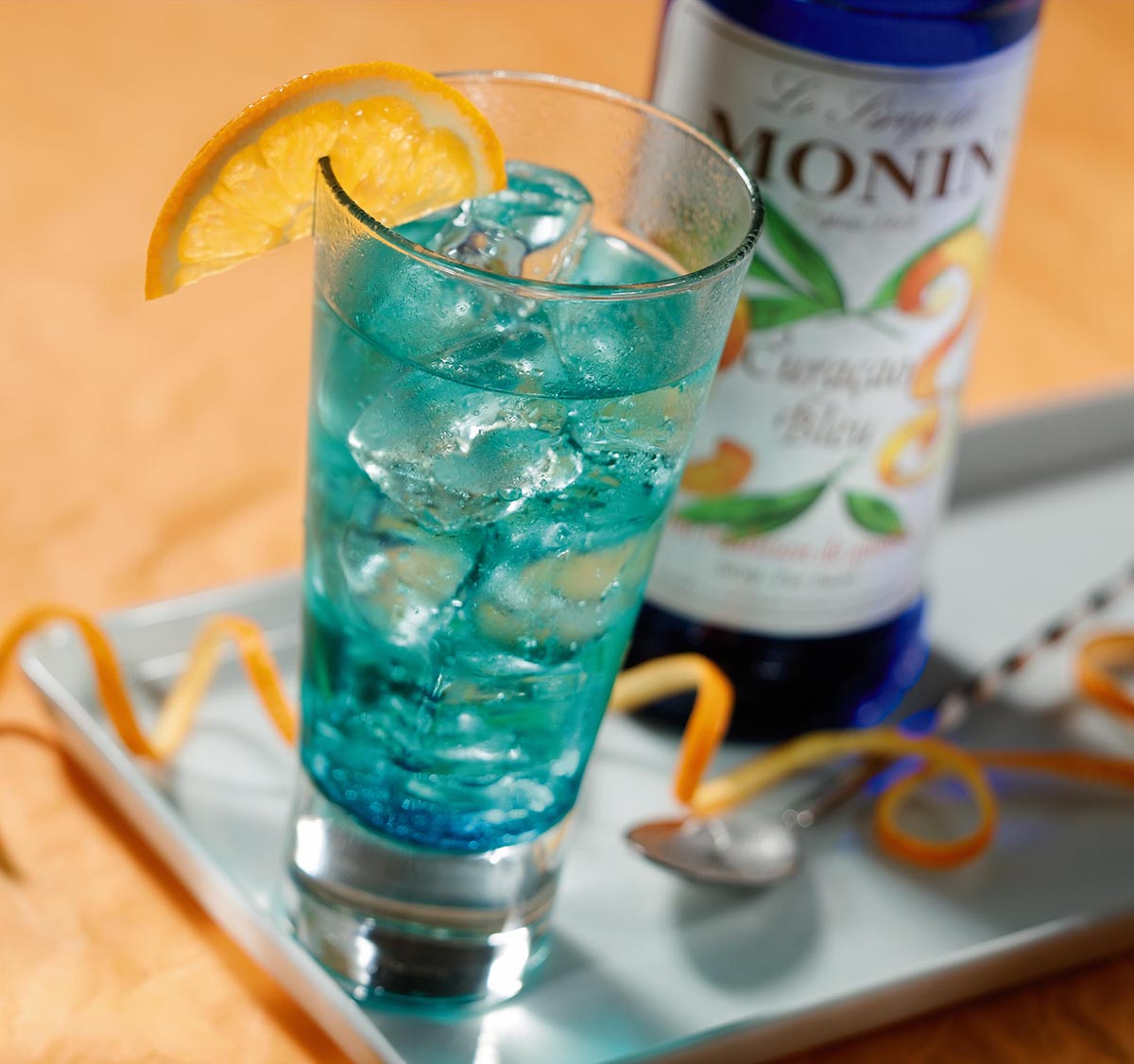 Blauer Cocktail mit Eiswürfeln und Orangenscheibe, Monin Blue Curaçao Sirup und Orangenschale.