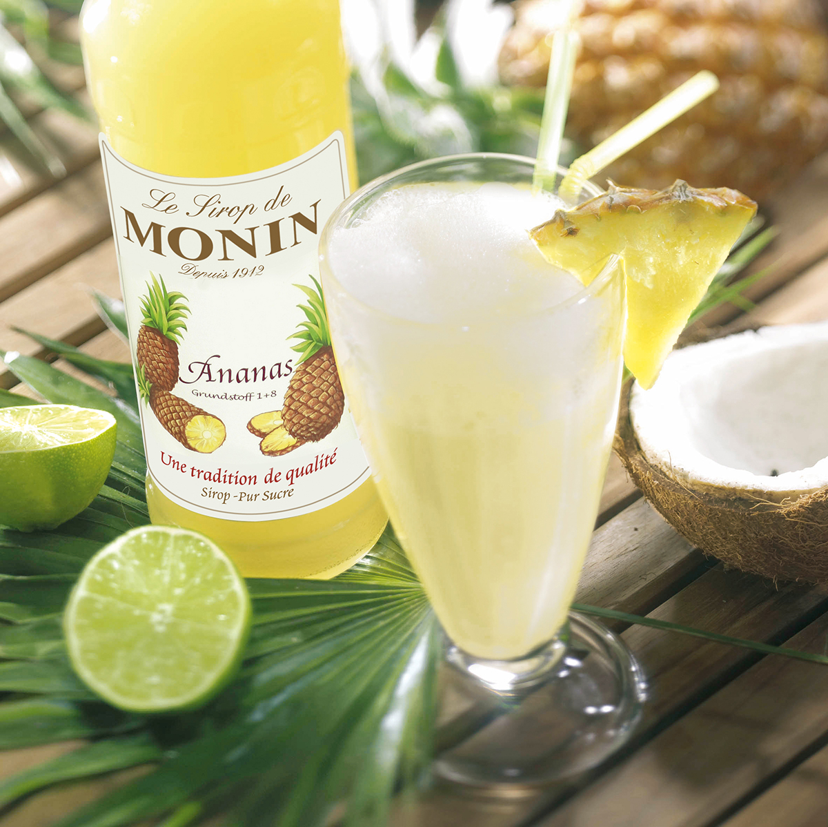 Cocktail mit Ananas, Limette und Monin-Sirupflasche. Kokosnuss und Palmenblatt in der Szene.
