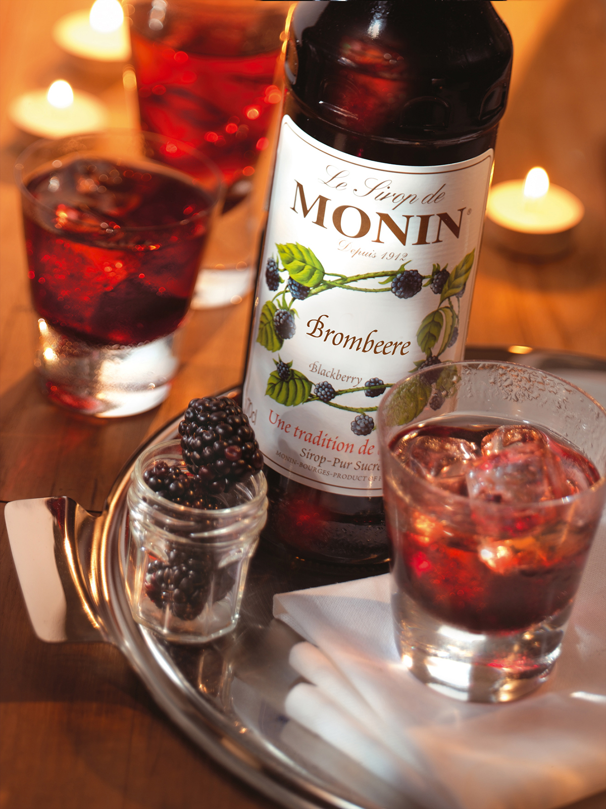 Brombeersirup, Gläser mit roten Getränken, Eis und Brombeeren auf einem Tablett, Kerzen.