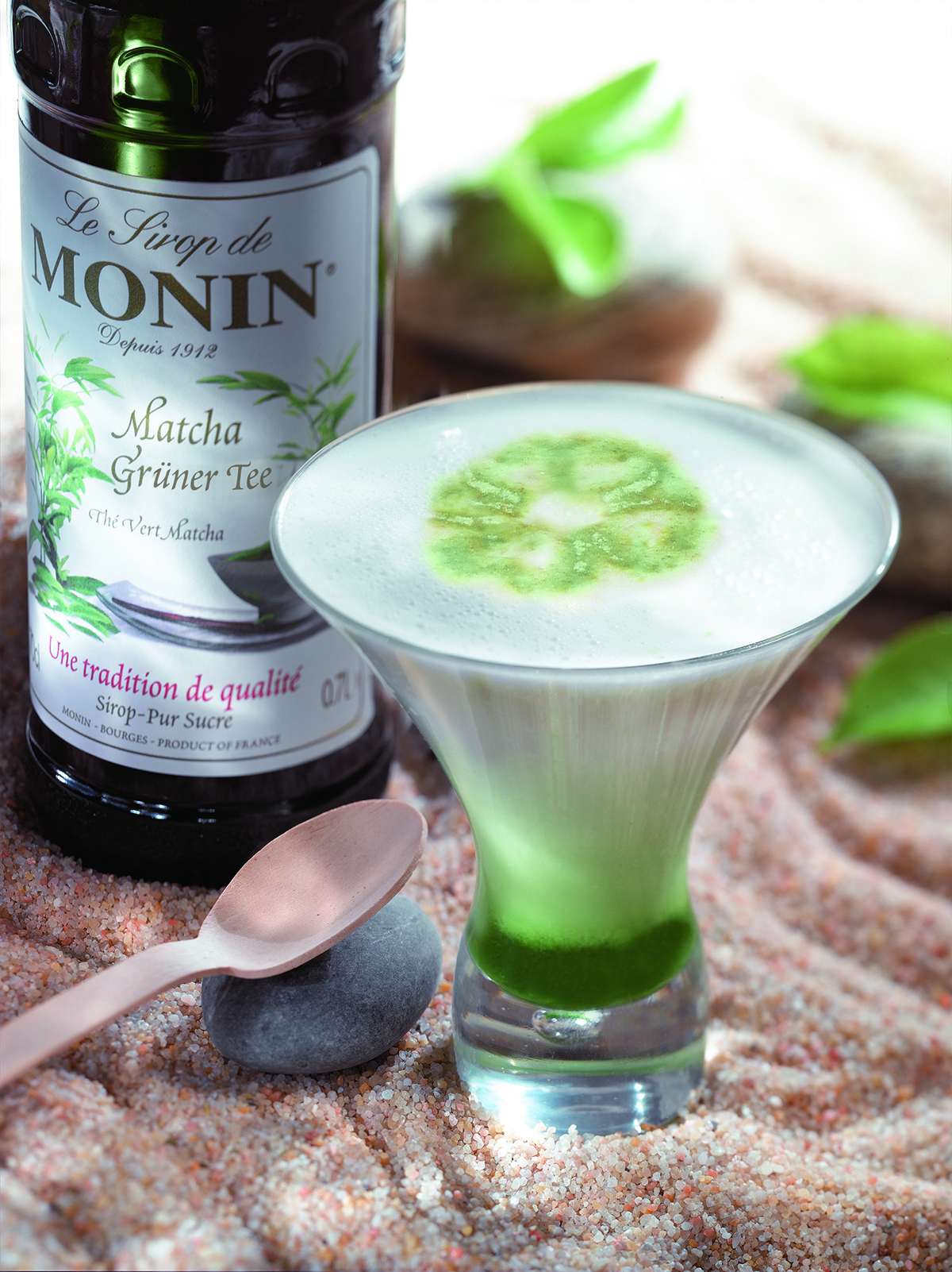 Matcha Latte mit Monin-Sirup, Holzlöffel und kleinen Steinen auf sandiger Oberfläche.