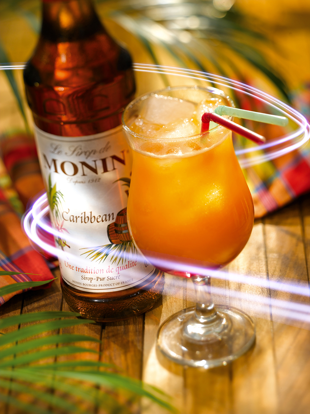 Cocktail im Glas mit Monin-Sirup und Eis, Strohhalmen und einer Flasche. Palmenblätter und ein kariertes Tuch daneben.