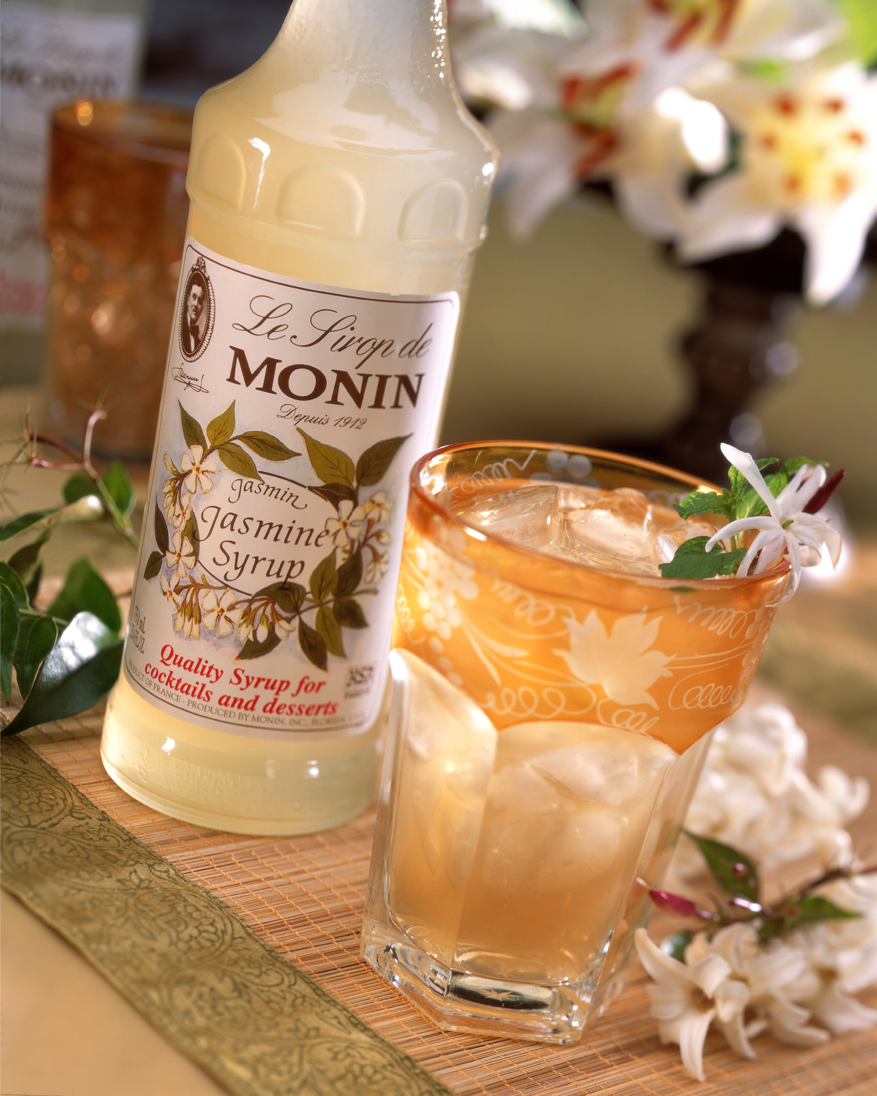 Eine Flasche Monin Jasmin-Sirup und ein Cocktailglas. Das Glas hat Eis und Blumen darin.