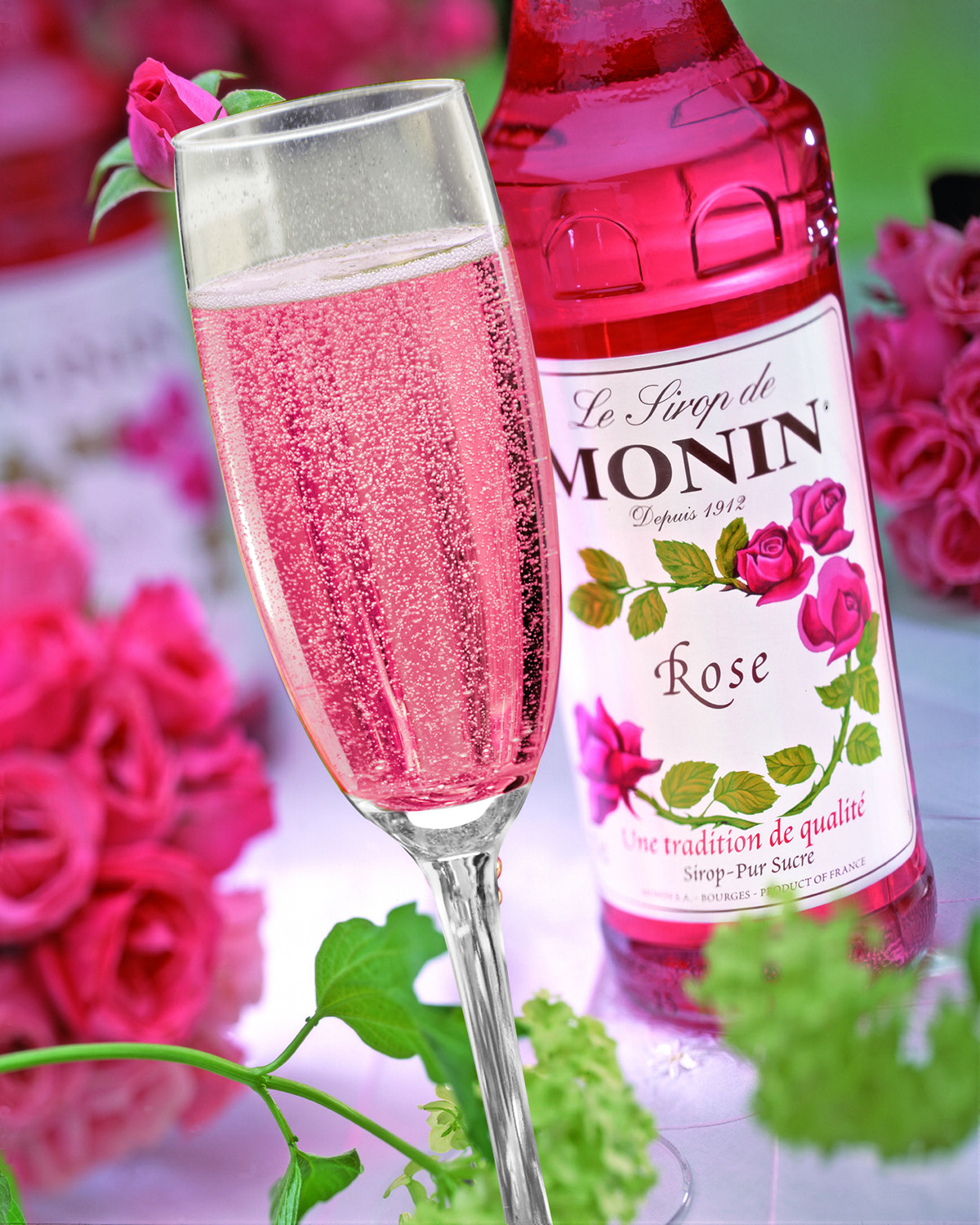 Rosafarbener Champagner mit Monin-Rosen-Sirup, rosa Rosen und floralem Hintergrund.