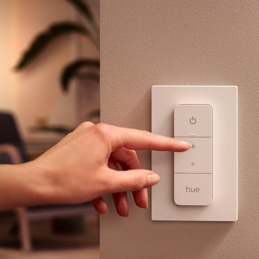 Une main appuie sur un bouton d'un interrupteur de lumière intelligent blanc monté sur un mur beige.
