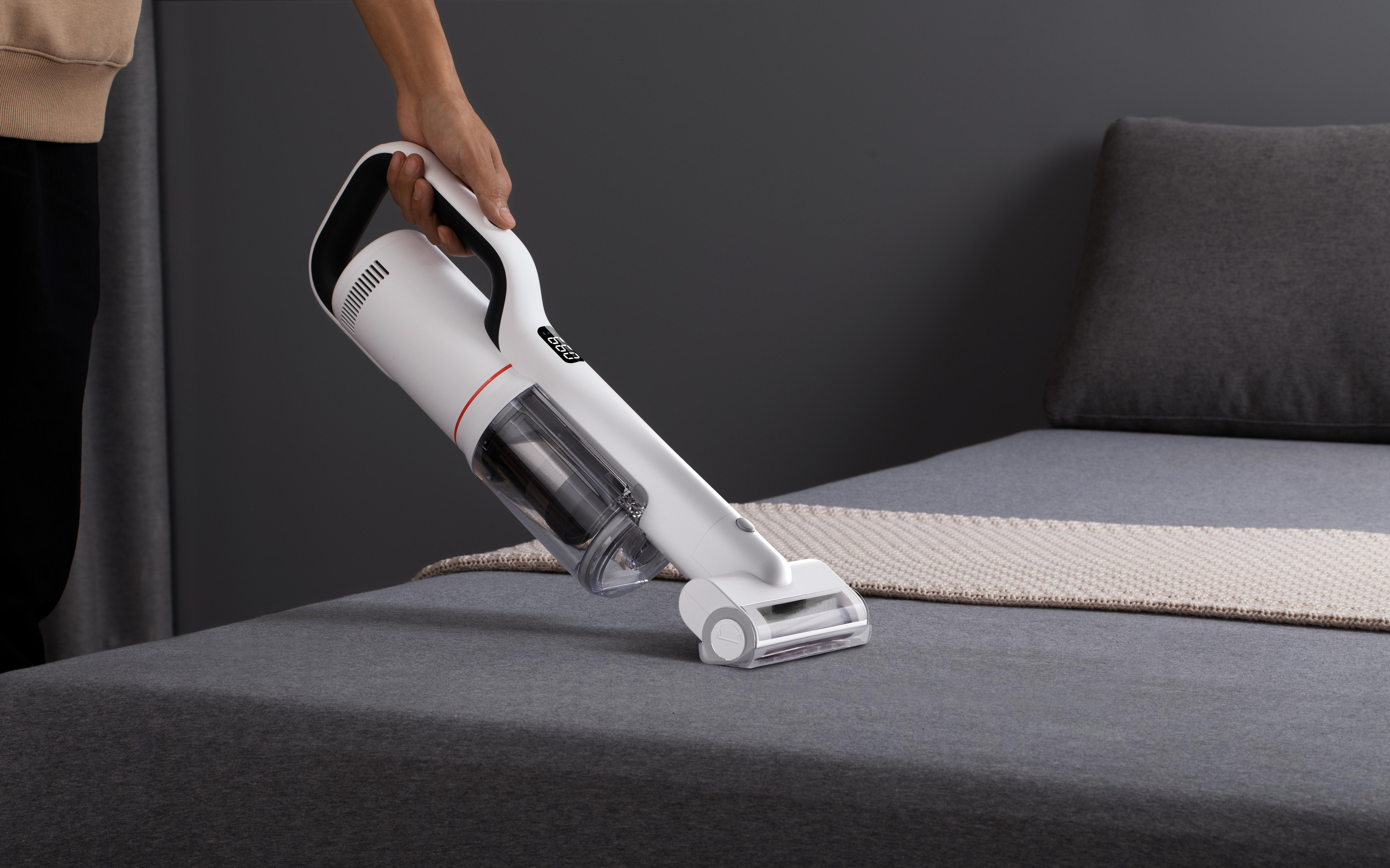 Person verwendet einen weißen Handstaubsauger auf einem grauen Bett.