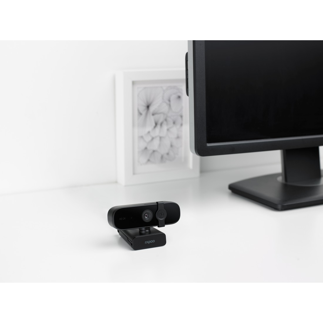 Schwarze Webcam auf einem Schreibtisch mit Monitor und gerahmtem Bild im Hintergrund.