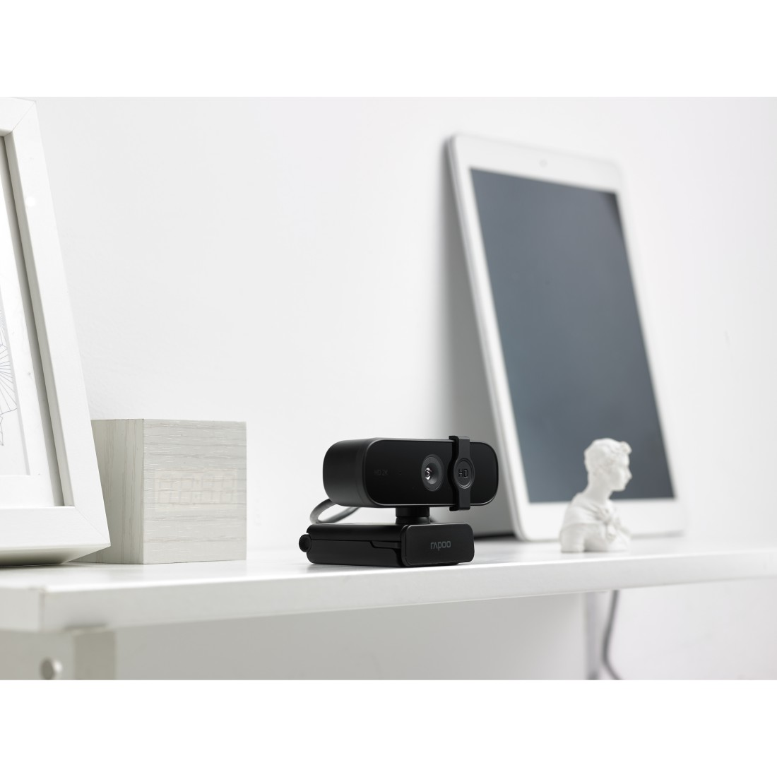 Eine schwarze Webcam auf einem weißen Regal mit Tablet und kleiner Statue.