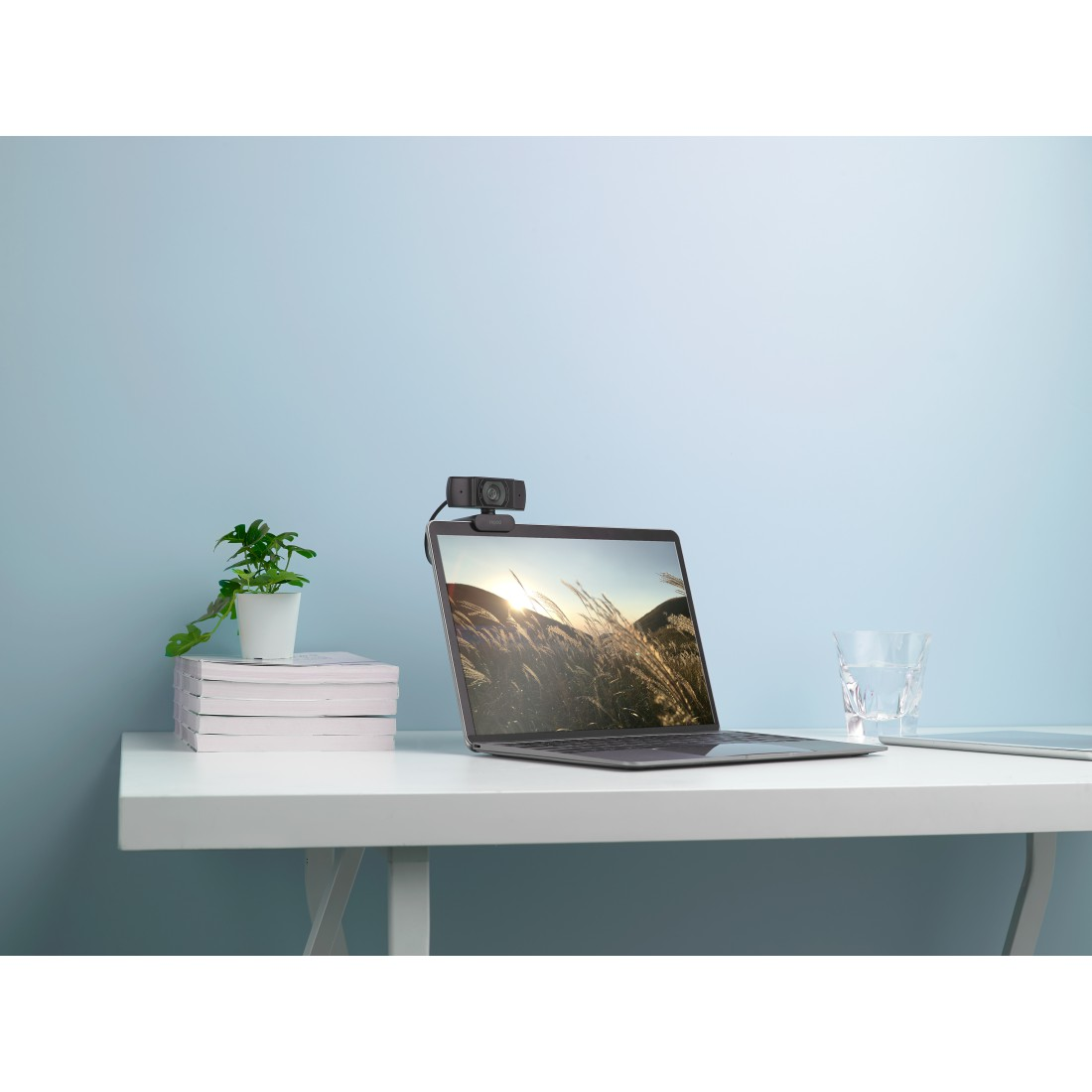 Laptop mit Webcam auf einem weißen Schreibtisch. Eine Pflanze, ein Wasserglas und Bücher stehen auf dem Schreibtisch. Hellblaue Wand im Hintergrund.