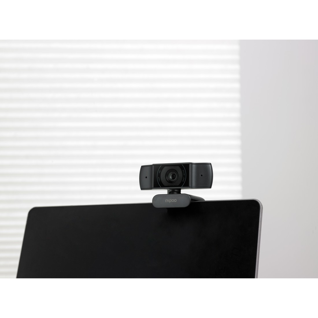 Eine schwarze Webcam sitzt auf einem Laptop-Bildschirm. Der Hintergrund zeigt eine weiße Wand mit horizontalen Jalousien. Die Webcam hat den Markennamen "rapoo".