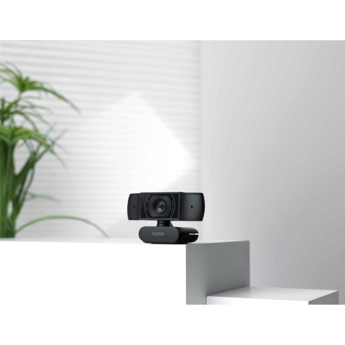 Eine schwarze Webcam steht auf einer weißen Oberfläche. Der Hintergrund ist weiß mit einer verschwommenen, hellen Jalousie und grünen Pflanzen.