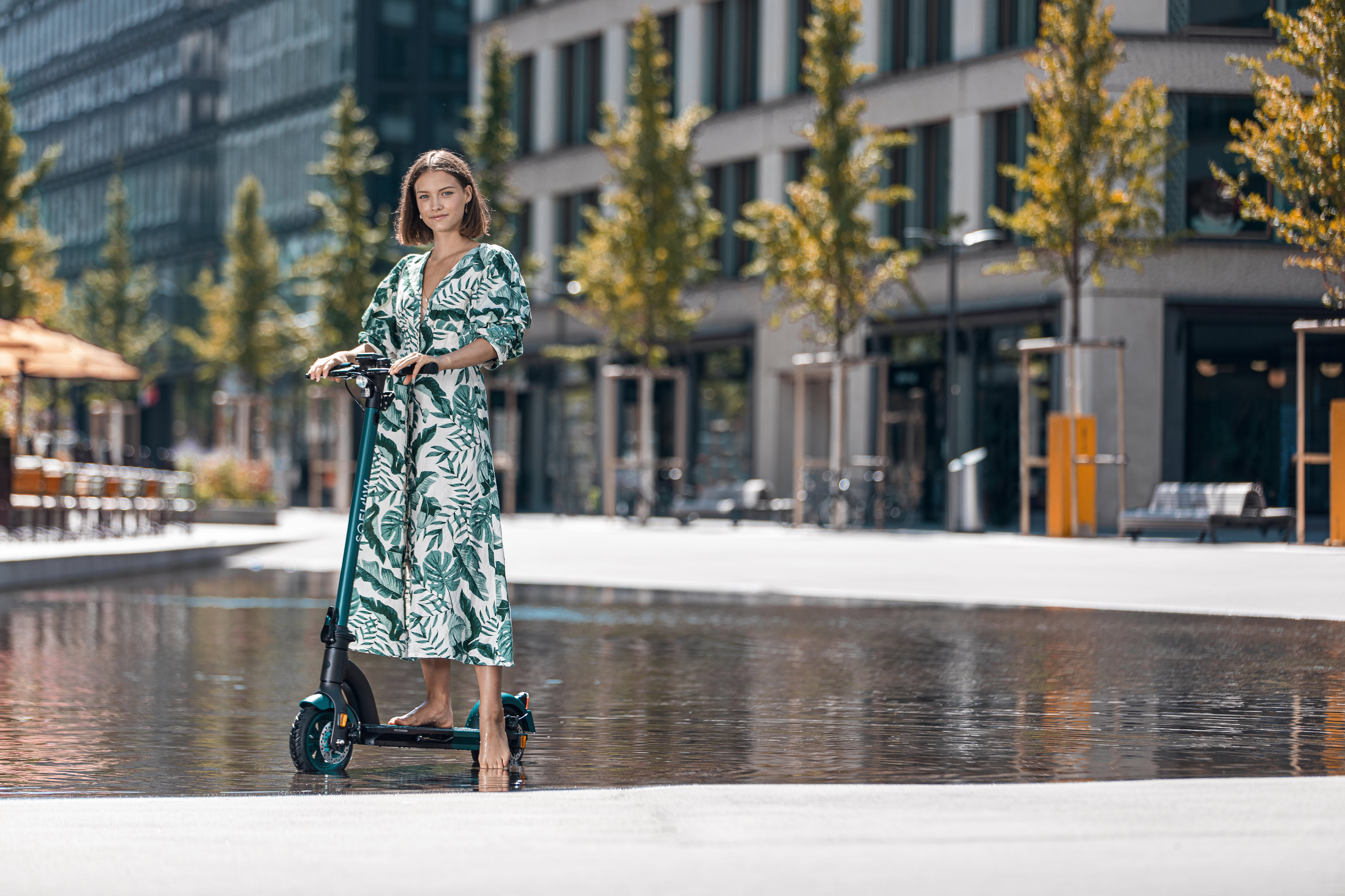 Eine Frau fährt einen grünen Elektroroller. Sie trägt ein weißes Kleid. Bäume und Gebäude im Hintergrund.