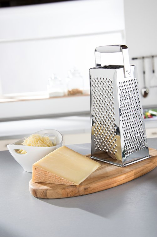 Käsereibe mit Käse, ein Stück Käse und eine Schüssel mit geriebenem Käse auf einem Holzbrett.