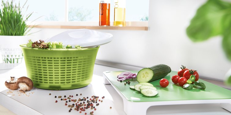 Küchenszene mit Salatschleuder, Gemüse und Zutaten auf einer weißen Arbeitsplatte.