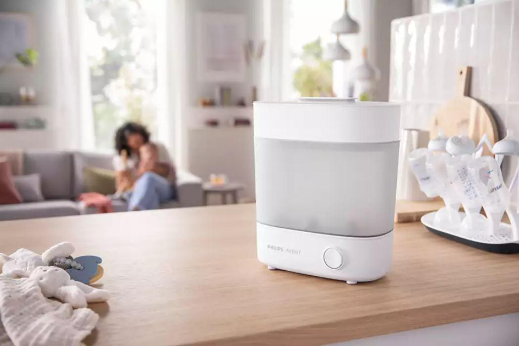 Ein weißer Babyflaschensterilisator mit Baby und Couch im Hintergrund.