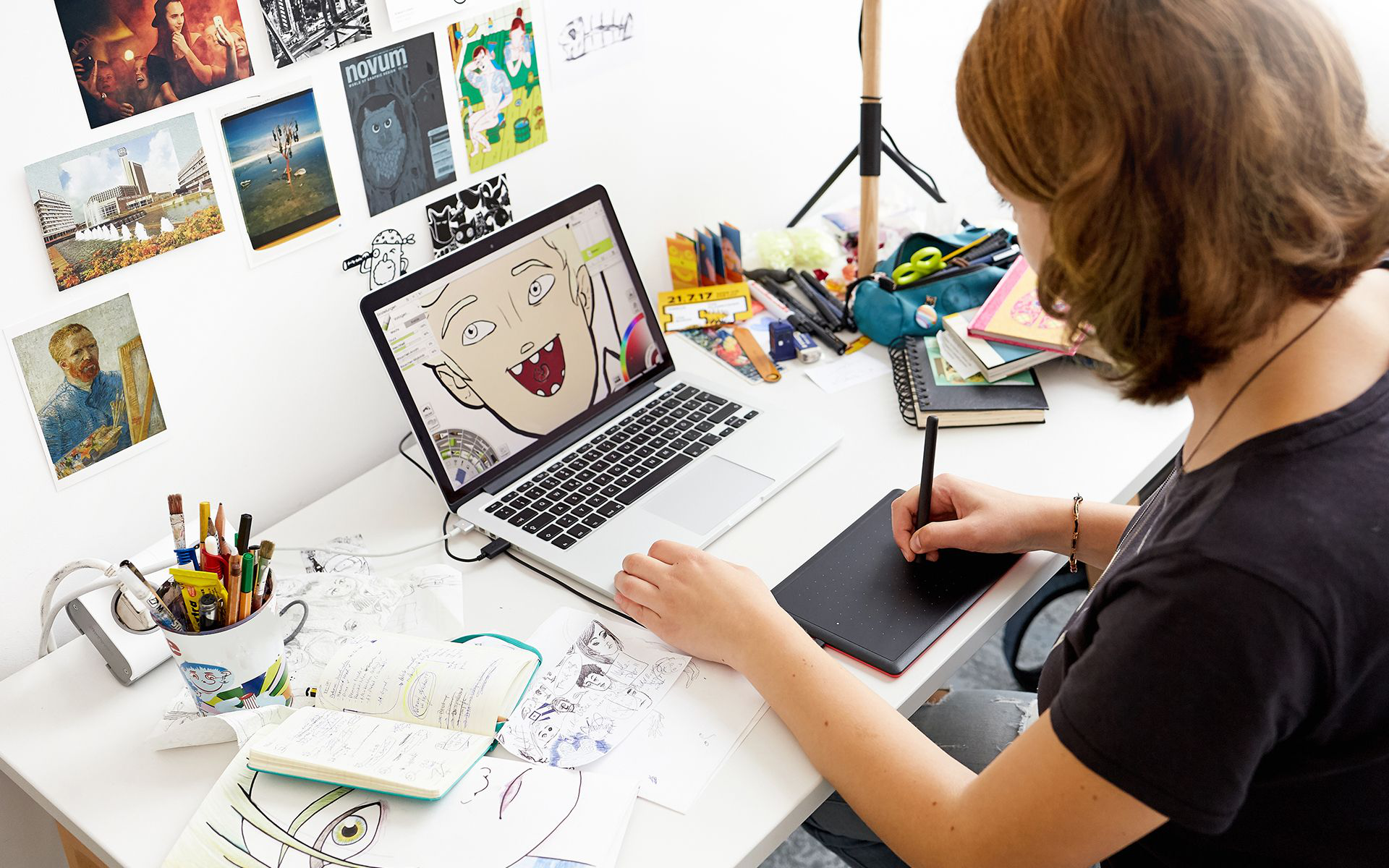 Donna che disegna su un laptop con una penna, circondata da materiali artistici.