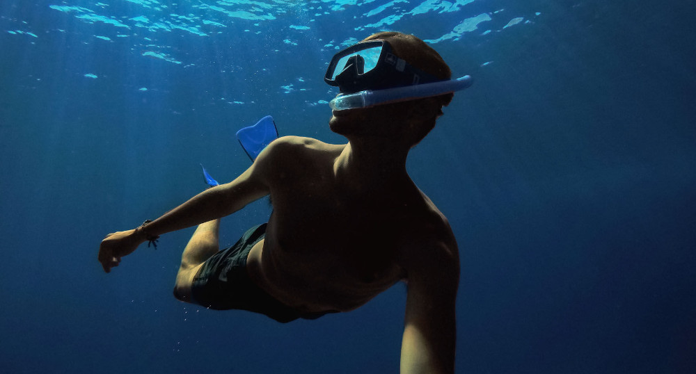 Une personne fait de la plongée avec tuba sous l'eau, avec un masque et des palmes. L'eau est bleue.