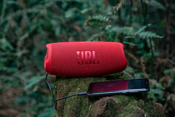 A red JBL speaker and a phone charging on a mossy log, set against a forest background.