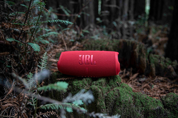 Red JBL speaker on a mossy log in a forest setting, surrounded by green foliage.