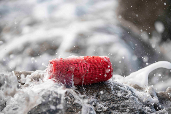 A red JBL speaker is hit by a wave on the beach.