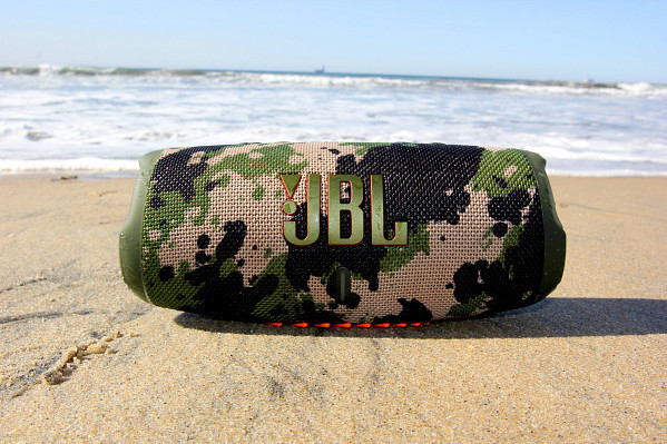 Camouflage JBL-luidspreker op een zandstrand met oceaangolven op de achtergrond.