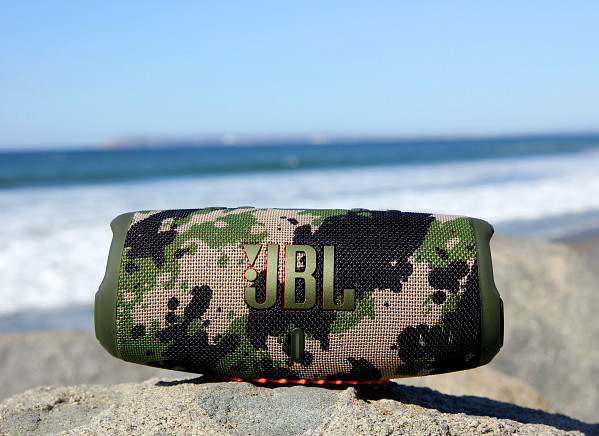 Camouflage JBL-luidspreker op een rots bij het strand, met blauwe lucht op de achtergrond.