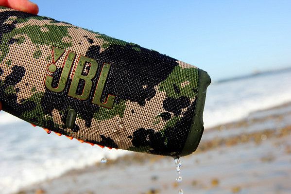 Een camouflage luidspreker met JBL-logo, waterdruppels en een strandachtergrond.