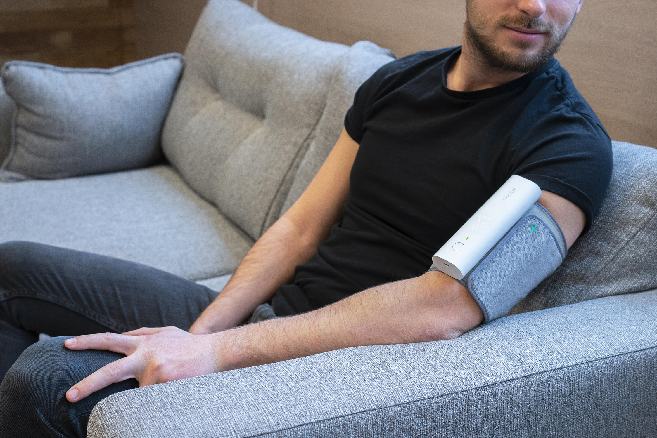 Man op een grijze bank heeft een bloeddrukmeter om zijn arm. Hij draagt een zwart shirt en een grijze broek. Het apparaat is wit.