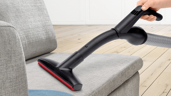 Person saugt mit einem schwarzen und roten Staubsaugerkopf ein graues Sofa ab.