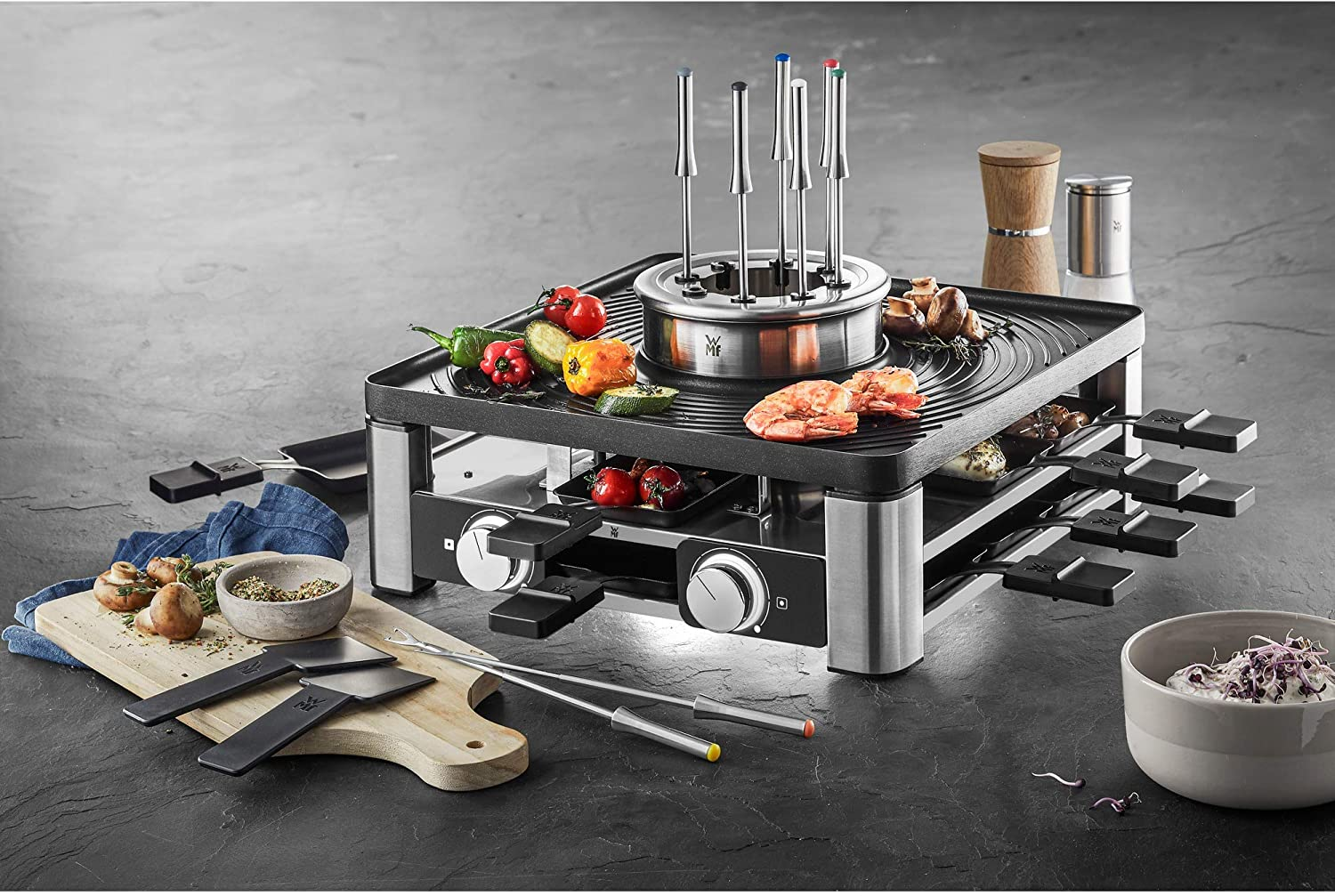 Ein Raclette-Grill mit verschiedenen Lebensmitteln, darunter Gemüse, Garnelen und Fleisch, auf schwarzer Oberfläche.