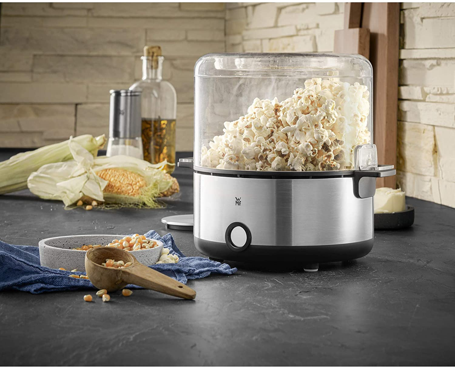 Popcornmaschine mit klarem Deckel, Edelstahlbasis und Körnern. Mais und Ölflaschen im Hintergrund.