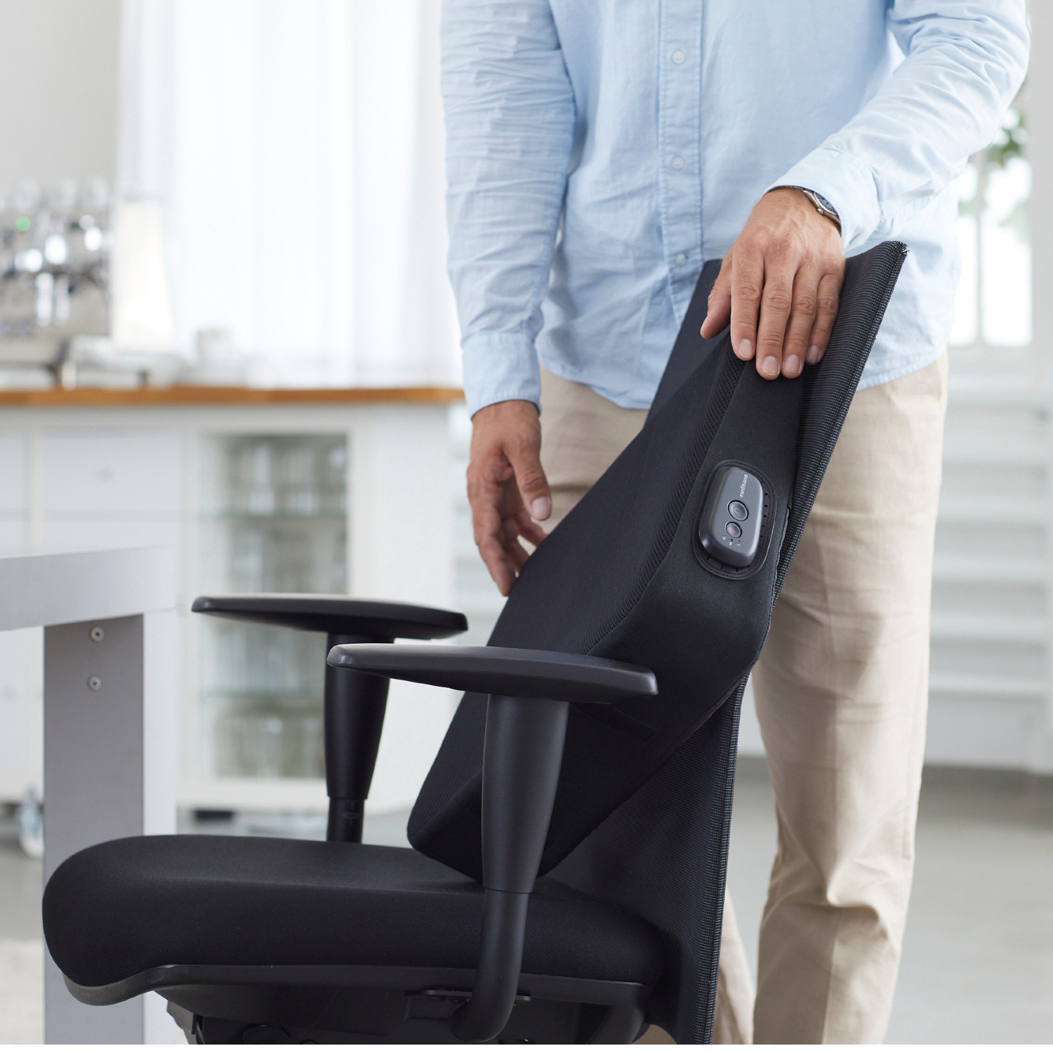 Un homme ajuste une chaise de bureau noire. Il porte une chemise bleu clair et un pantalon beige. L'arrière-plan montre une cuisine lumineuse.