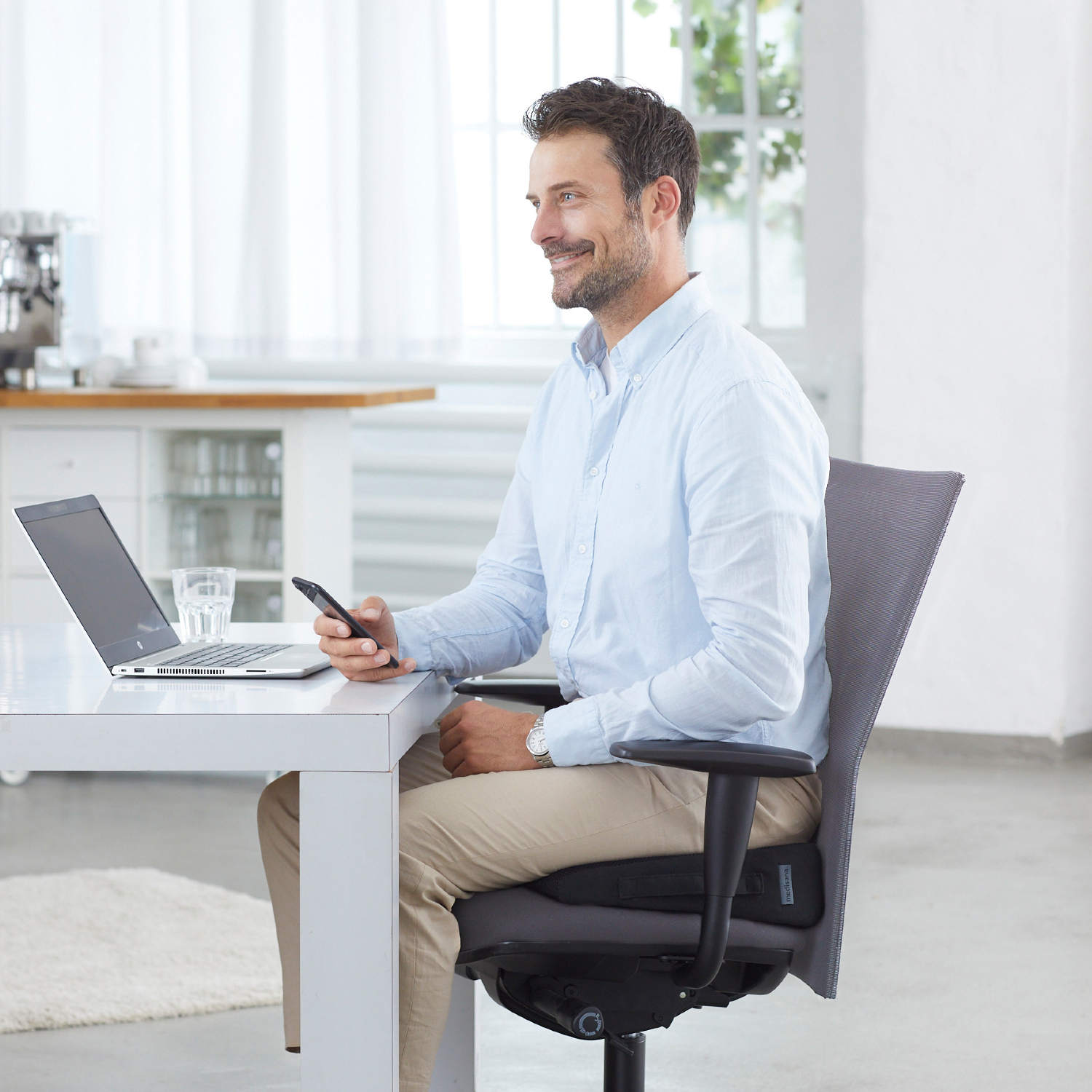 Un homme est assis à une table avec un ordinateur portable et un téléphone, souriant. Il porte une chemise bleu clair et un pantalon kaki. Il y a une fenêtre.