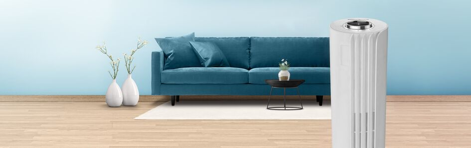 A living room scene: blue sofa, flowers in vases, and a white air purifier on a wooden floor.