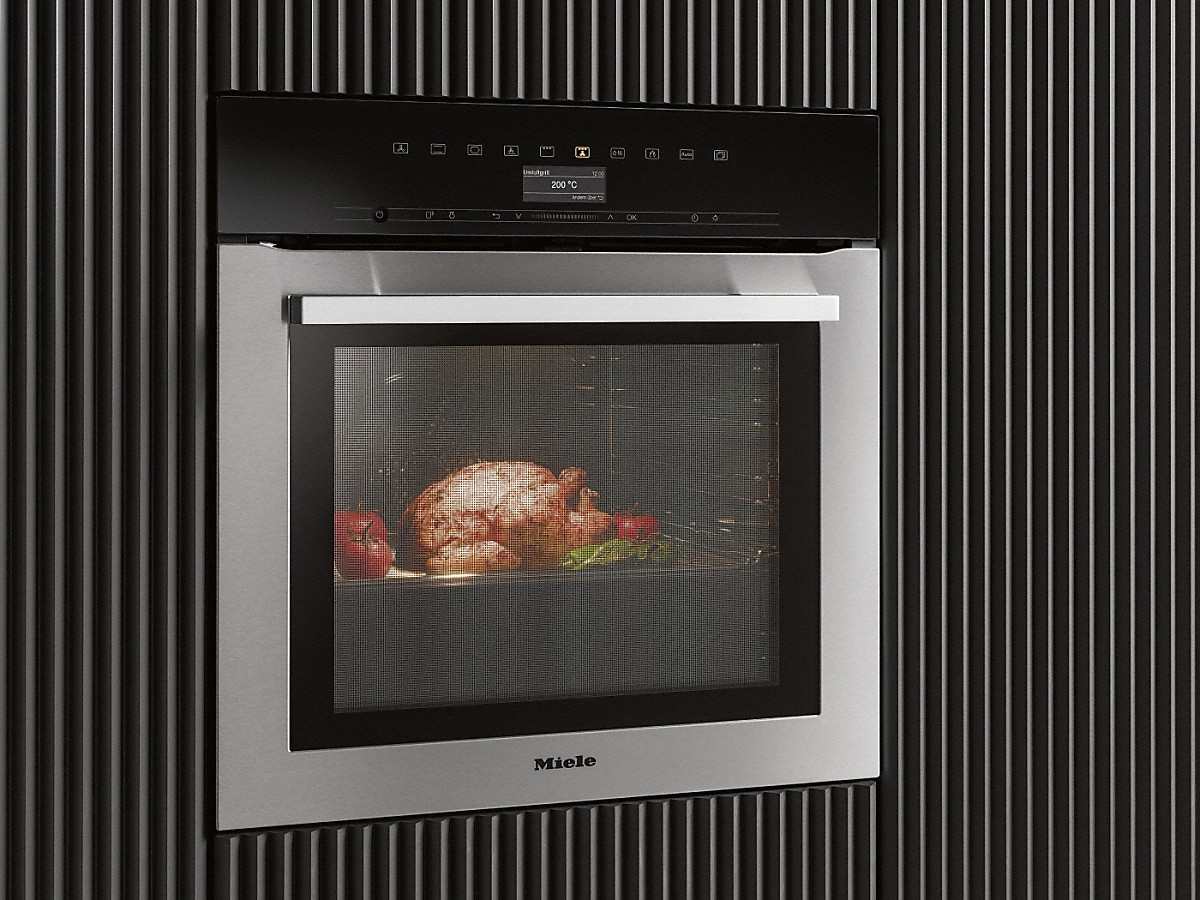 Ein Edelstahlbackofen mit einem Huhn darin. Der Hintergrund ist eine schwarze Wand mit vertikalen Linien.