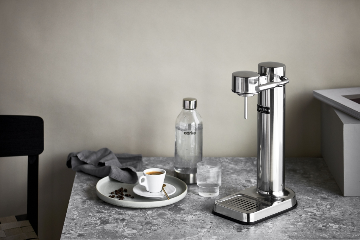 Kaffeemaschine aus Edelstahl mit einer Glaswasserflasche, Tasse mit Kaffee, einem Teller mit Bohnen, auf einer hellgrauen Theke.