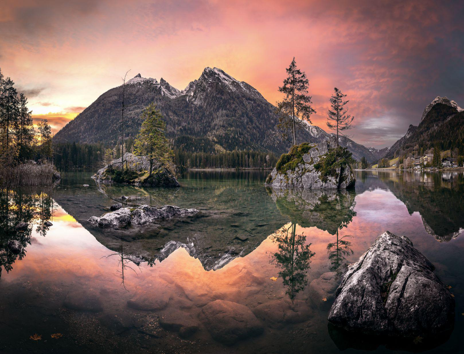 Eine malerische Landschaft mit See, der Berge, Bäume und einen farbenfrohen Sonnenuntergang spiegelt.