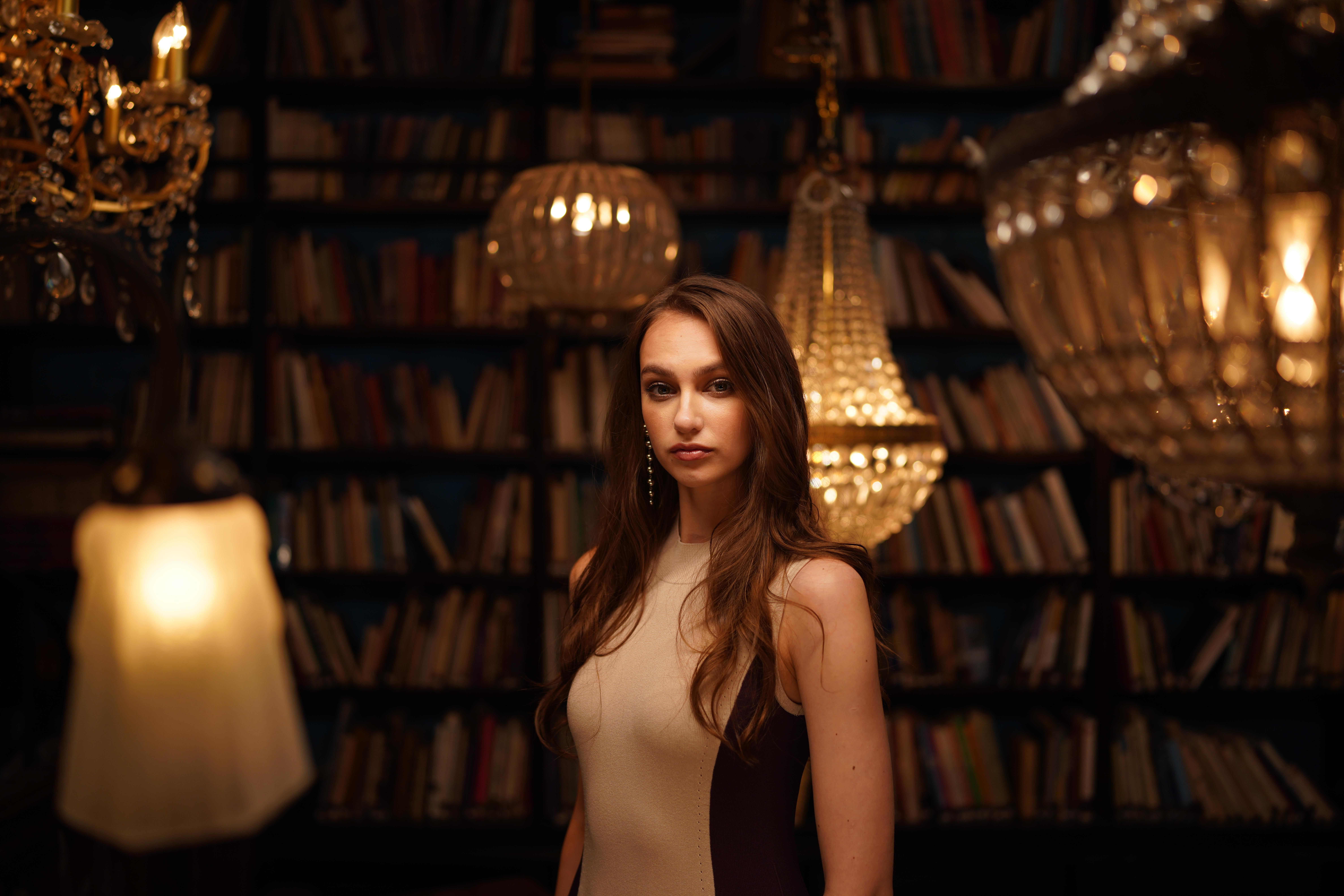 Eine Frau in einem beige-burgunderfarbenen Kleid steht in einer Bibliothek mit Kronleuchtern und Büchern.