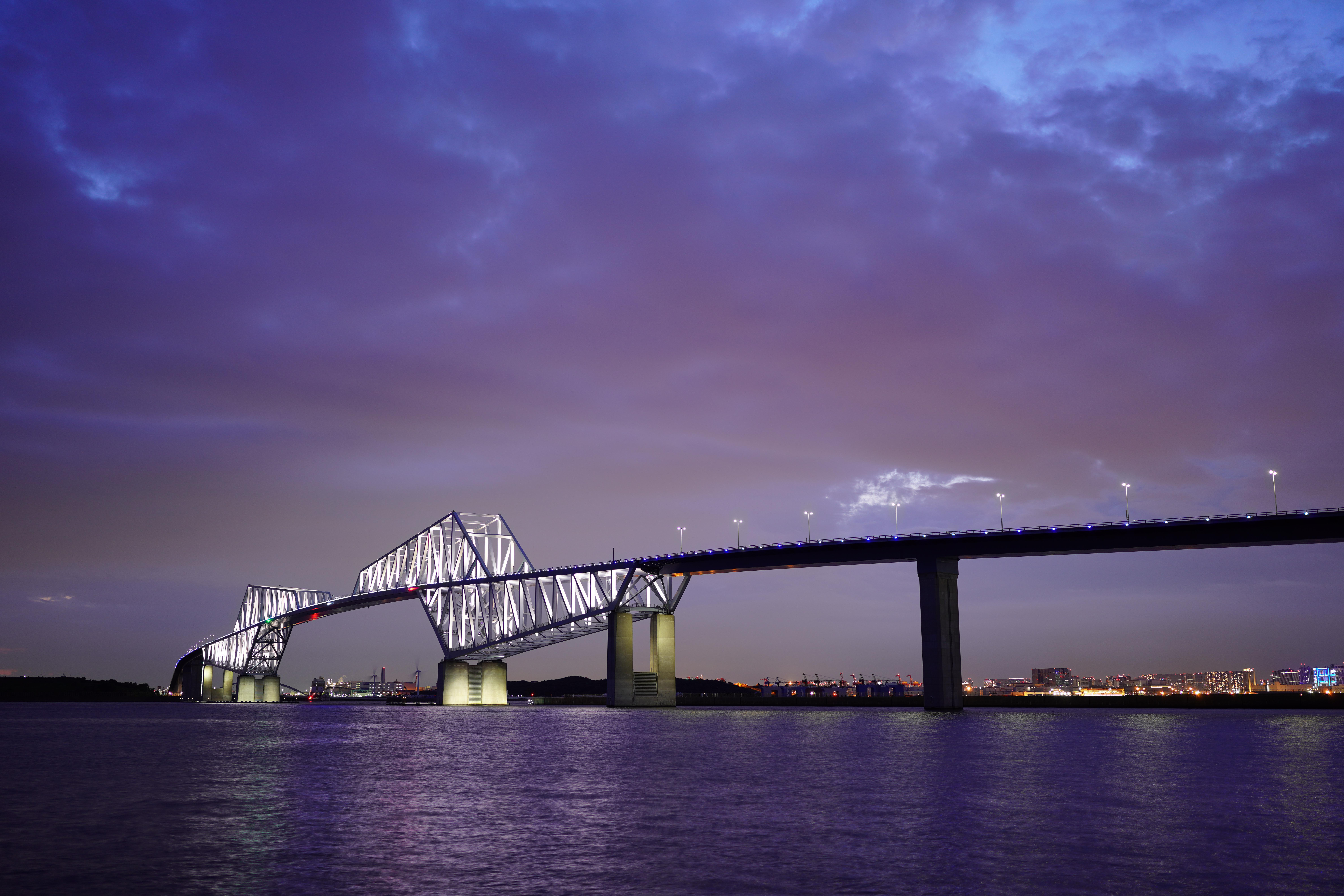 Nachtansicht einer beleuchteten Brücke über Wasser. Der Himmel ist dunkelblau mit Wolken. Stadtlichter sind in der Ferne sichtbar.